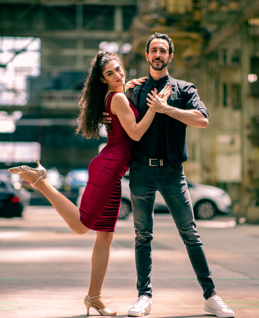Eine Frau im roten Kleid und ein Mann im schwarzen Hemd posieren verspielt in einer industriellen Umgebung.
